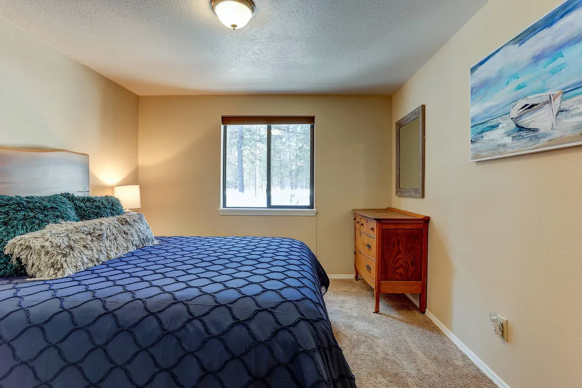 A bright, inviting bedroom with a bed covered in a quilted blue cover, a window streaming in natural light, a wooden dresser, and a charming painting of a boat on the wall. The space feels restful and inviting for guests.