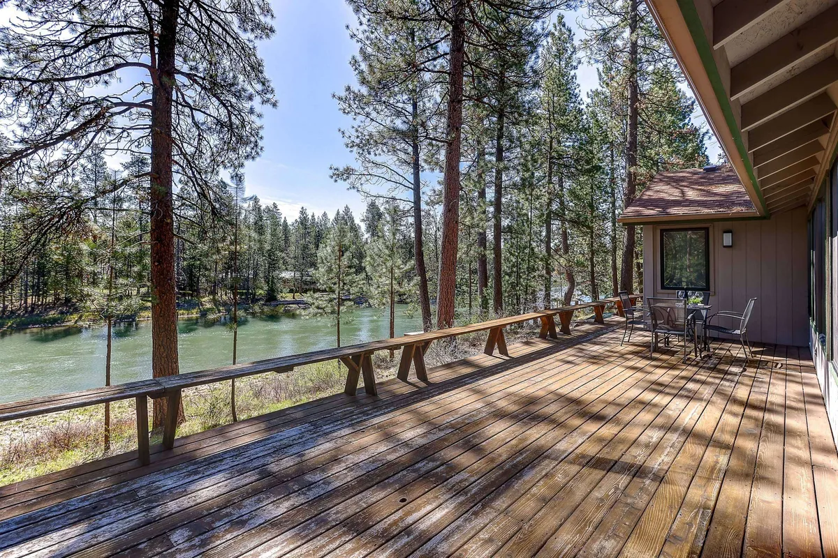 A spacious wooden deck overlooks the serene Deschutes River, framed by lush trees and offering comfortable outdoor seating. The tranquil water and natural setting invite guests to relax, dine, and observe wildlife or sunsets from the deck.
