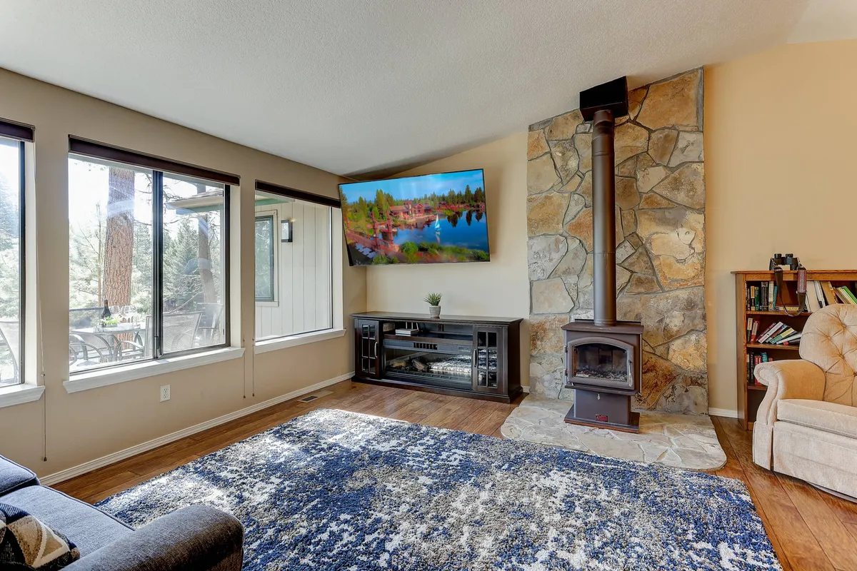 A spacious living room featuring a stone fireplace as its centerpiece, a plush sofa, a decorative rug, and a wall of large windows that flood the space with natural light and showcase tranquil views of the river outside.