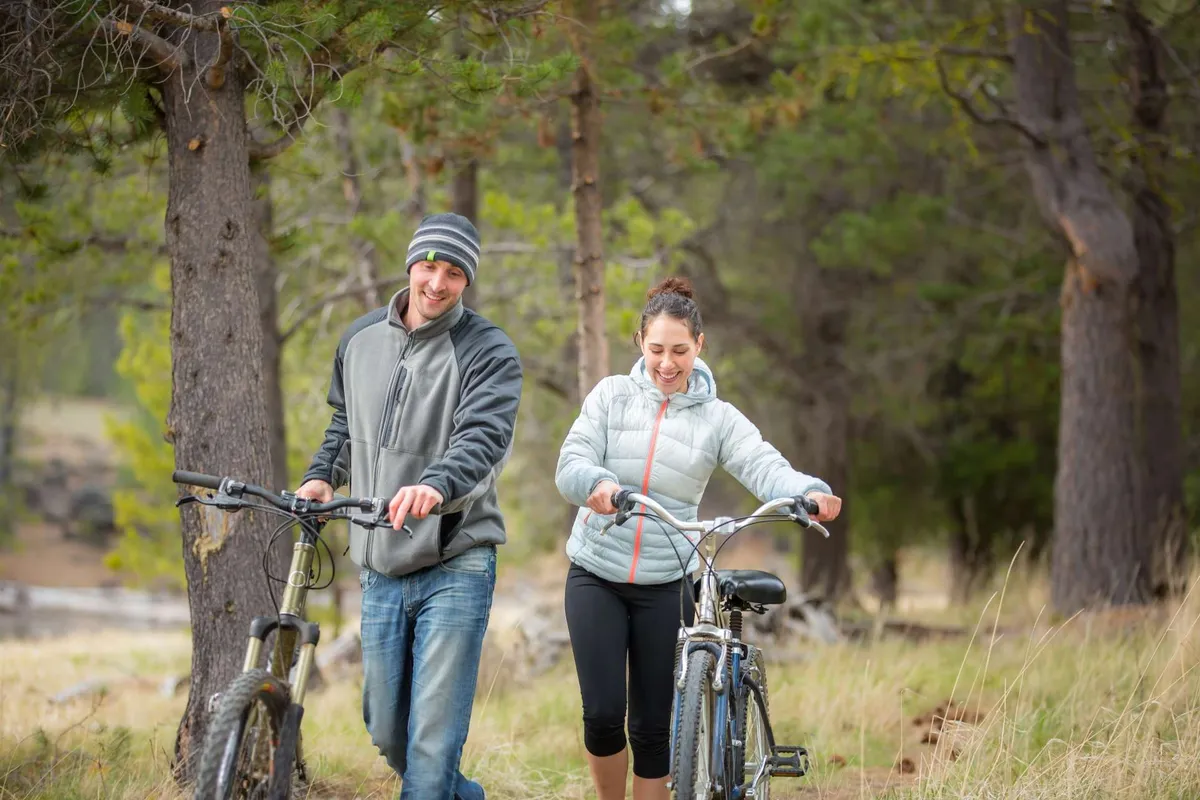 Two cyclists, accompanied by tall trees and lush natural landscape, walk with their bikes along a path. The image radiates joy and adventure, with a sense of togetherness and exploration.