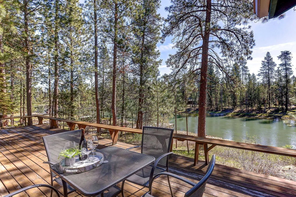 The image showcases an inviting outdoor dining table set on a spacious deck, overlooking the serene Deschutes River. Towering pine trees frame the view, and the soft evening light hints at the promise of unforgettable sunset gatherings. The natural surroundings evoke a sense of peace and seclusion, perfect for unwinding or watching wildlife.