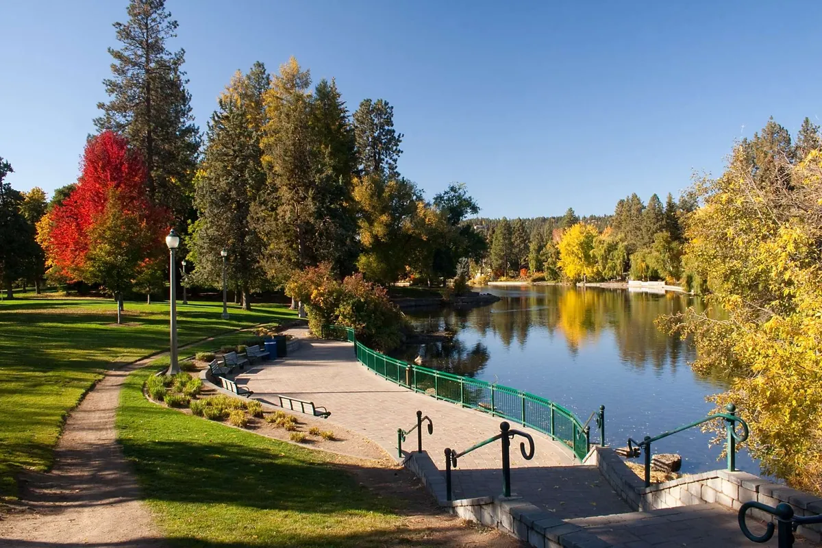 A calm lake is bordered by colorful autumn trees, with a park pathway and green railing inviting exploration. The natural setting is serene, offering opportunities for walking, picnicking, and wildlife watching.