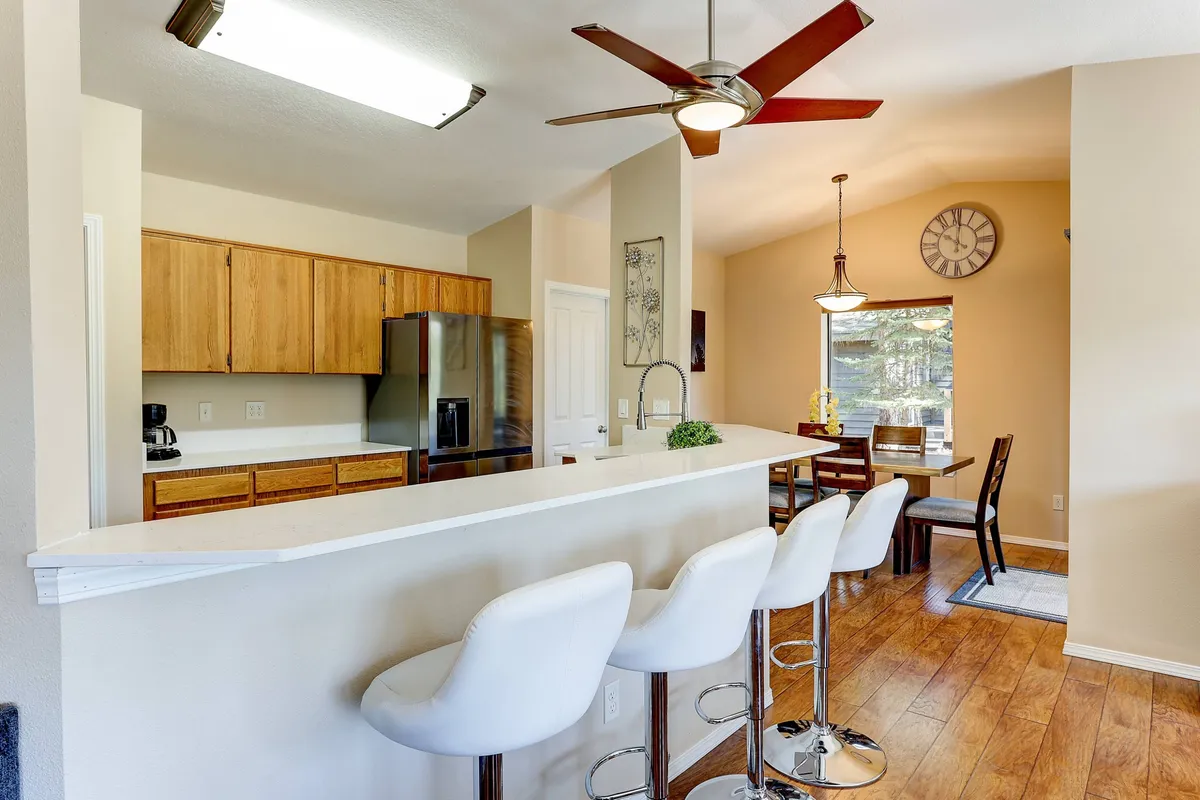 A wide-angle view of the open-plan area that includes a kitchen island with bar stools, a dining area, and a large window streaming in natural light. The space is designed for family meals, entertaining, and seamless connection between kitchen and living.