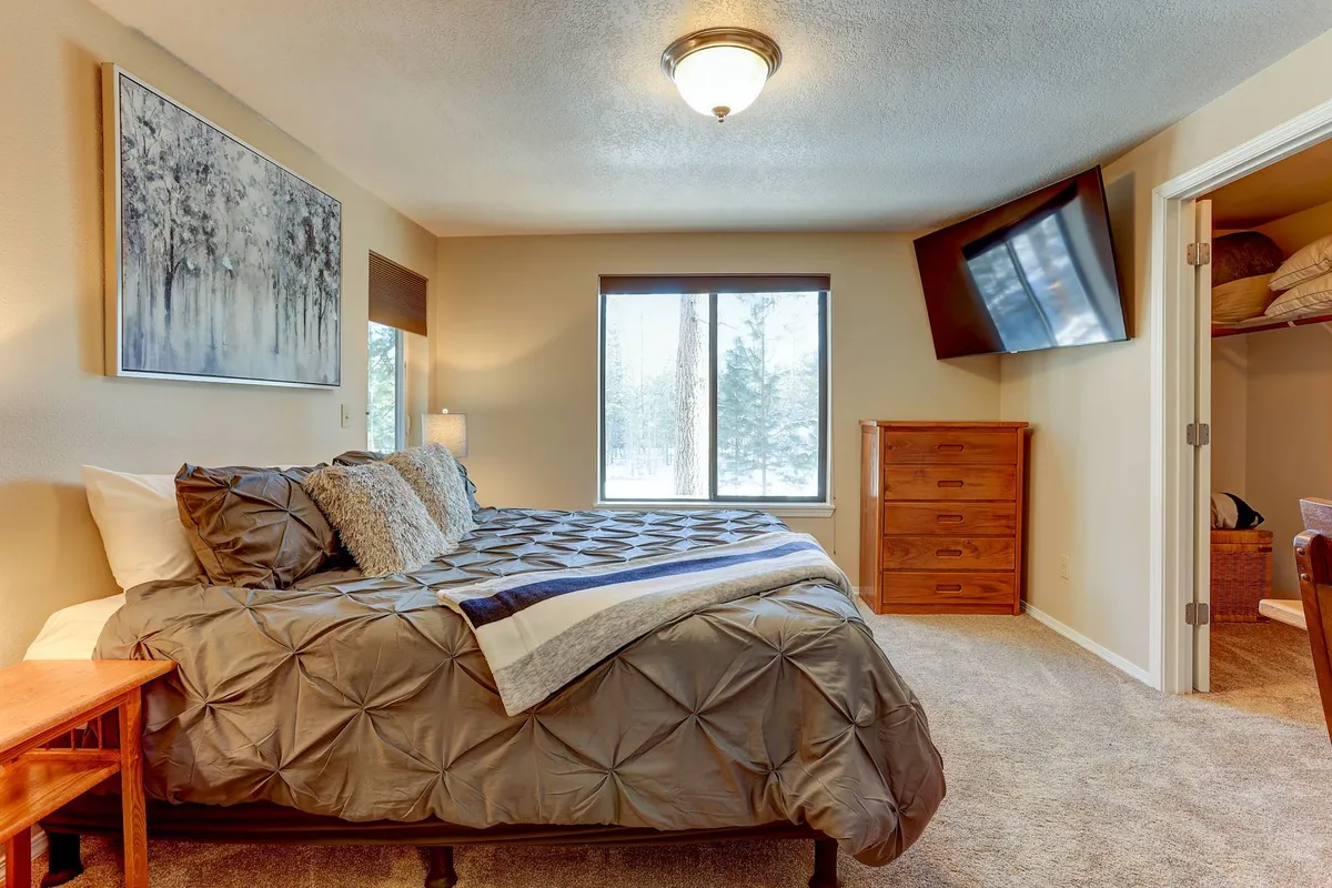 A warmly lit bedroom featuring a comfortable bed with decorative pillows, a large window allowing ample natural light, a dresser, and a TV mounted on the wall. The view outside the window suggests tranquility and connection to nature.