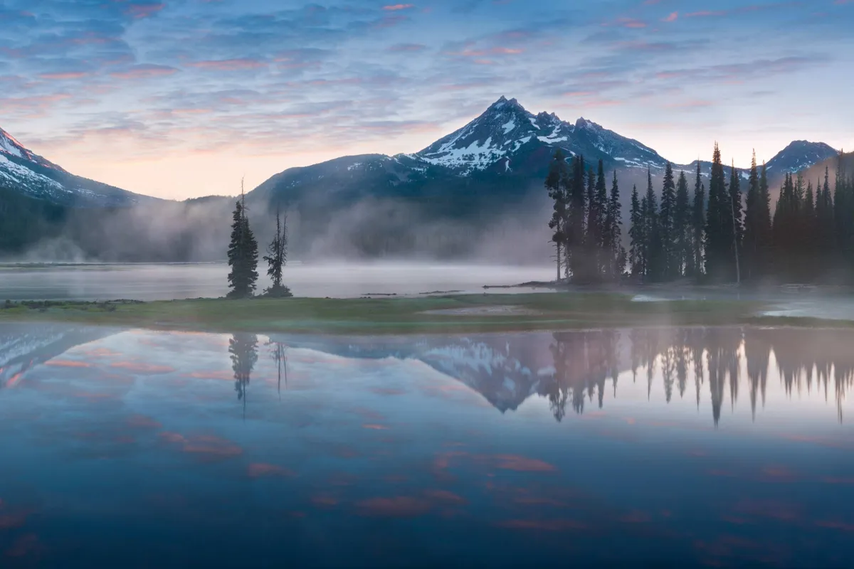 A tranquil view of a reflective mountain lake at sunrise, surrounded by misty pine trees and distant peaks. The water’s surface mirrors the soft colors of the morning sky, evoking peace and a sense of natural beauty.