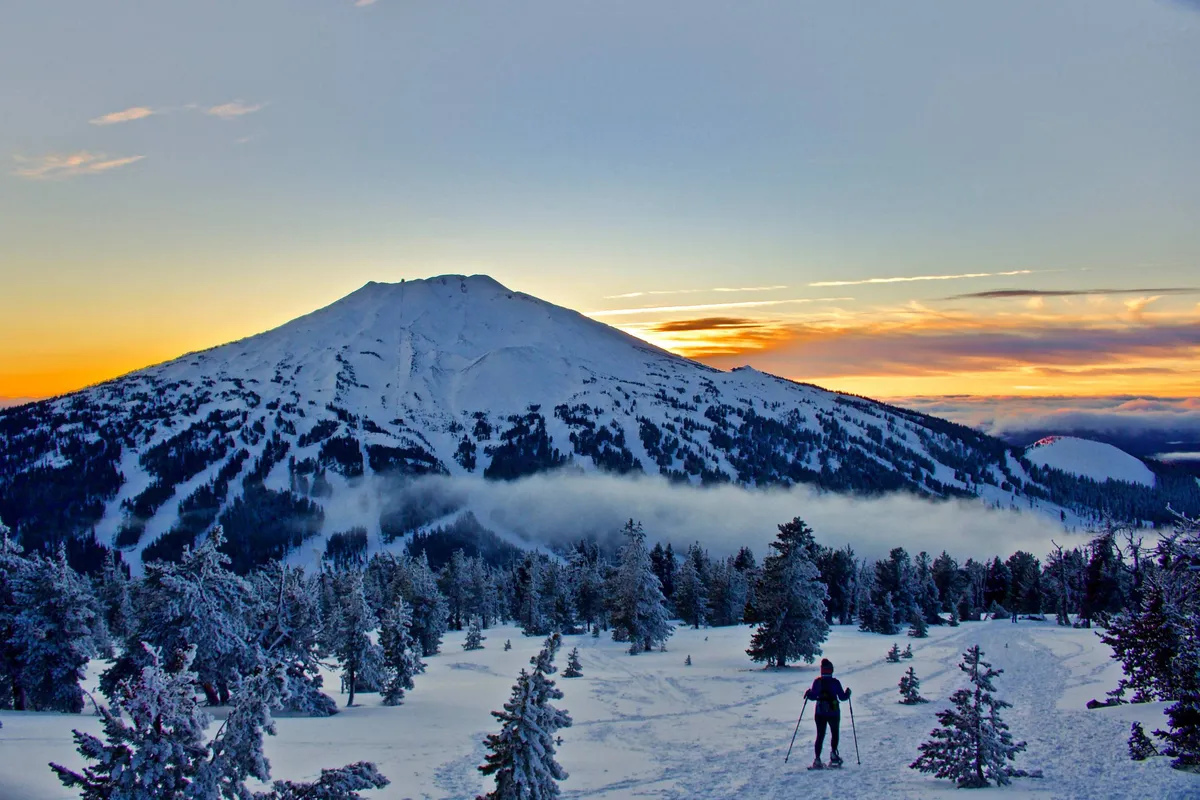 A dramatic view of a snow-covered mountain with a skier racing down the slope at sunset, surrounded by snowy trees. The scene captures the thrill of winter sports and the beauty of nature in colder months.